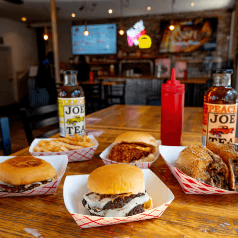 Halal smashburgers, ribeye cheesesteak, loaded fries and chicken sandwich on a wooden table at Cousin's Burger