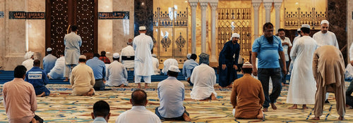 Men gathered for congregational Friday prayer inside a mosque with a patterned carpet