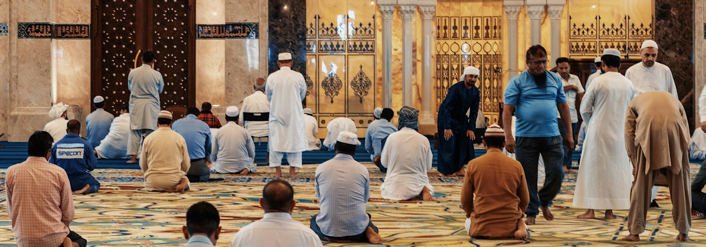 Men gathered for congregational Friday prayer inside a mosque with a patterned carpet