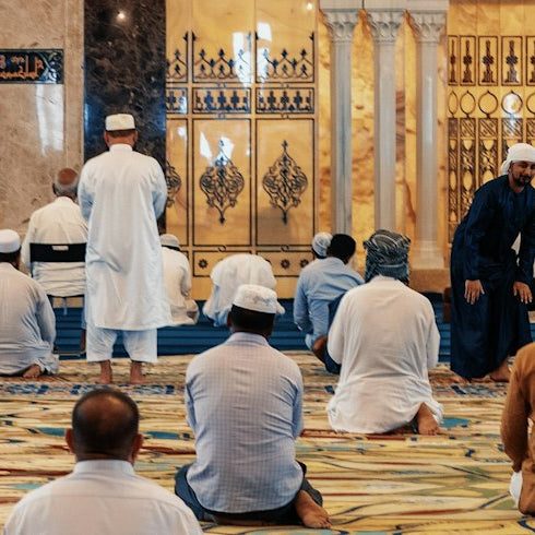 Men gathered for congregational Friday prayer inside a mosque with a patterned carpet