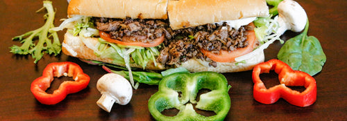 A loaded steak sandwich on a wooden table surrounded by fresh peppers and mushrooms