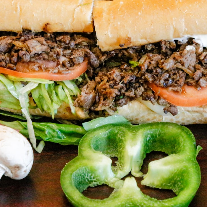 A loaded steak sandwich on a wooden table surrounded by fresh peppers and mushrooms